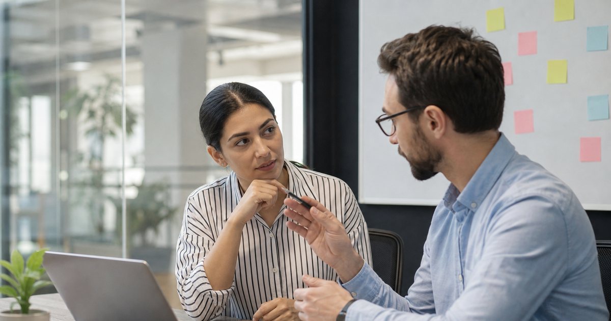 Two coworkers at a small startup office table talking over an open laptop, one holding a pen and gesturing, a whiteboard with colored sticky notes in the background, natural daylight