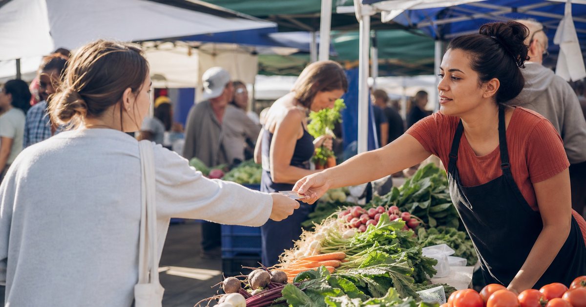 A vendor at a farmers market handing a small paper card to a customer across a table of fresh produce, morning sunlight, other shoppers browsing nearby stalls